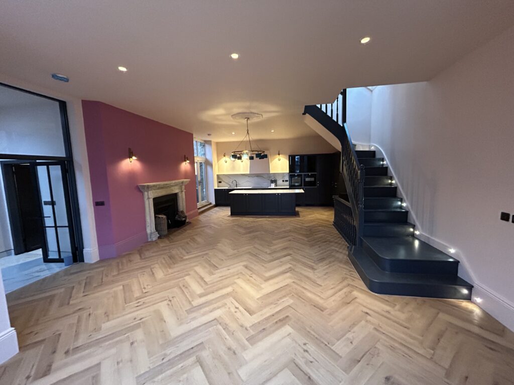 Open-plan living space in a renovated house, dusky pink feature wall and a dark open-tread staircase with lit treads, herringbone floor running through to the kitchen beyond