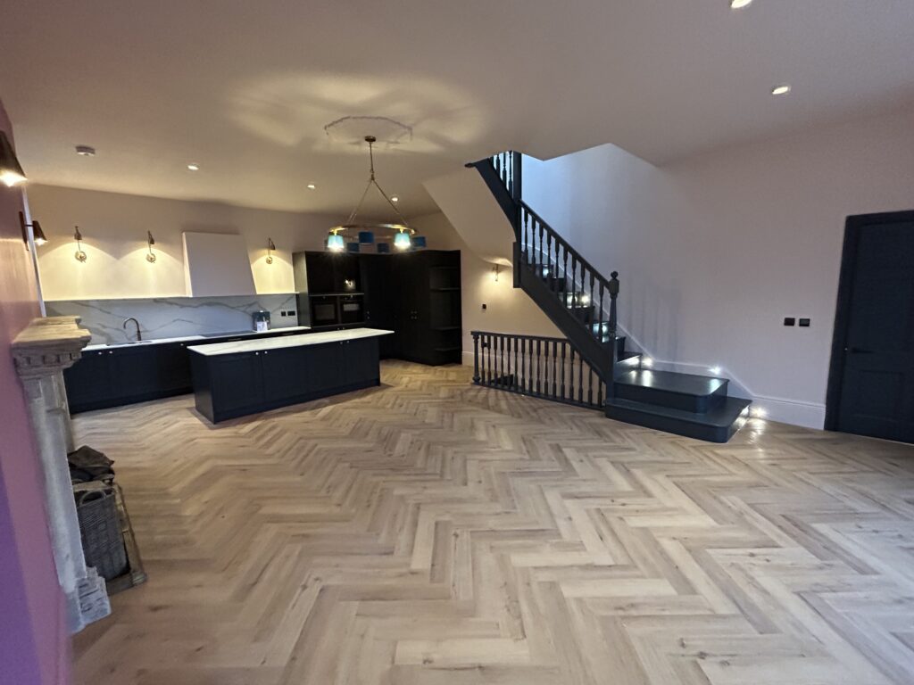 Renovated open-plan kitchen and living space, dark handleless units and island lit from above, herringbone floor and an open-tread staircase rising to the first floor
