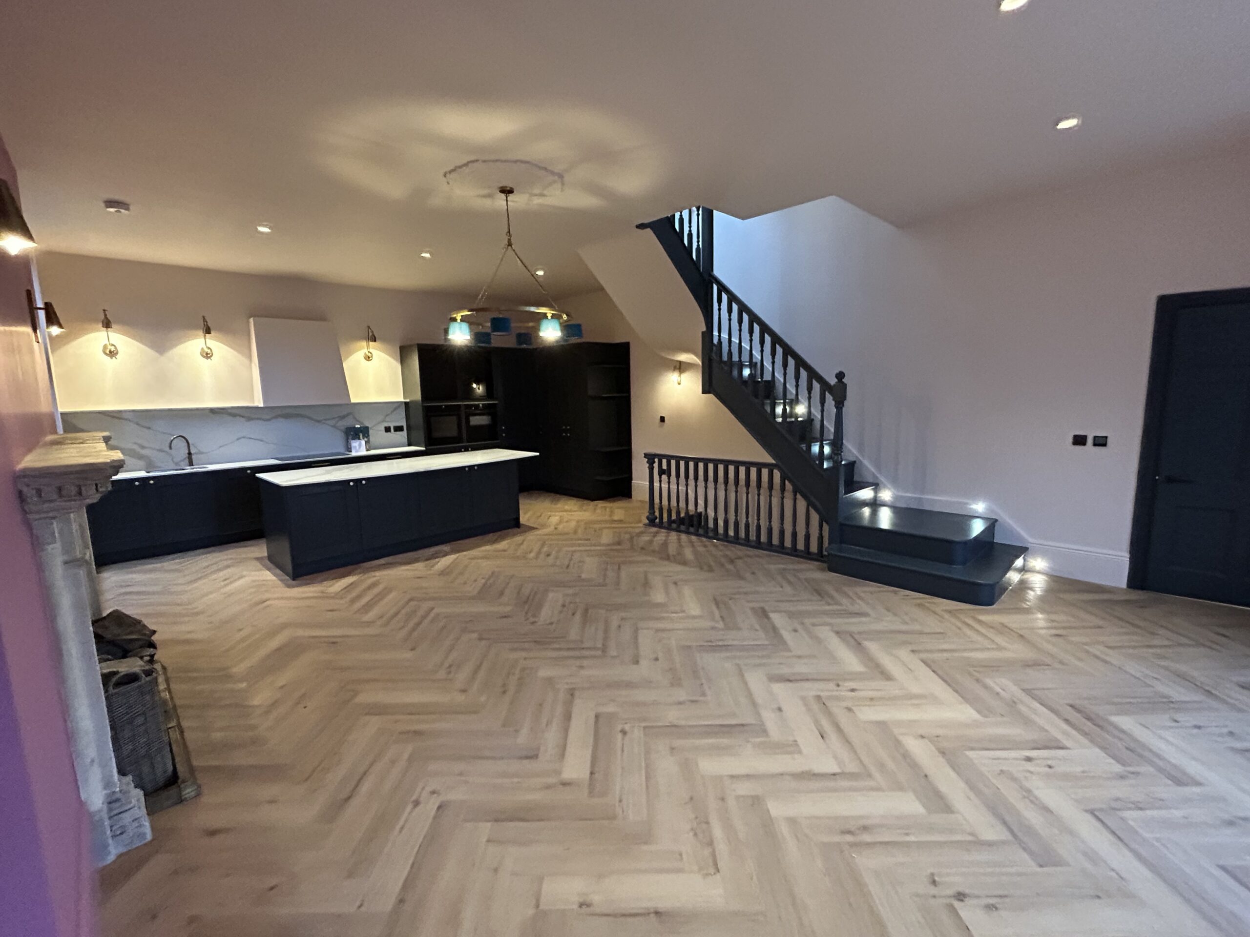 Renovated open-plan kitchen and living space, dark handleless units and island lit from above, herringbone floor and an open-tread staircase rising to the first floor