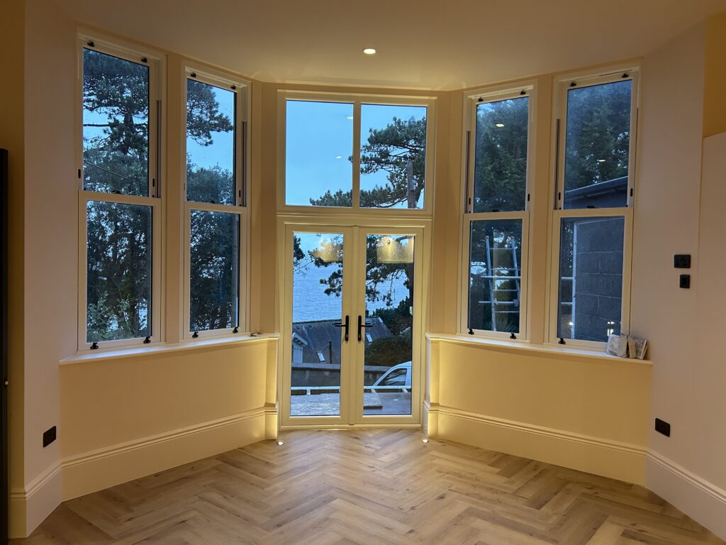 Restored bay window in a renovated period room, three tall sashes opening onto mature trees, herringbone floor and freshly plastered walls