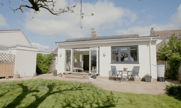 Rear of a white rendered bungalow with a single-storey extension, sliding doors open onto a paved patio and lawn