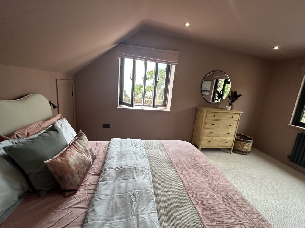 Loft bedroom with dusky pink walls, a window seat outlook and a wooden chest of drawers