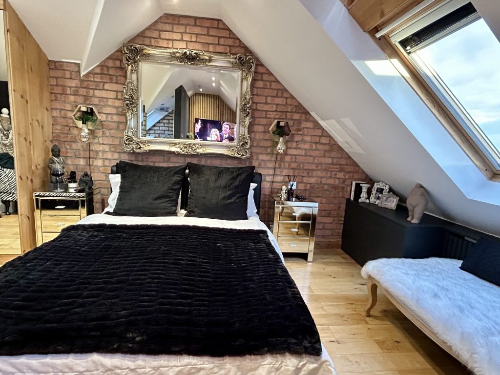 Loft bedroom with a double bed against an exposed brick chimney breast, a large gilt mirror, oak slatted panelling and a wide roof window.