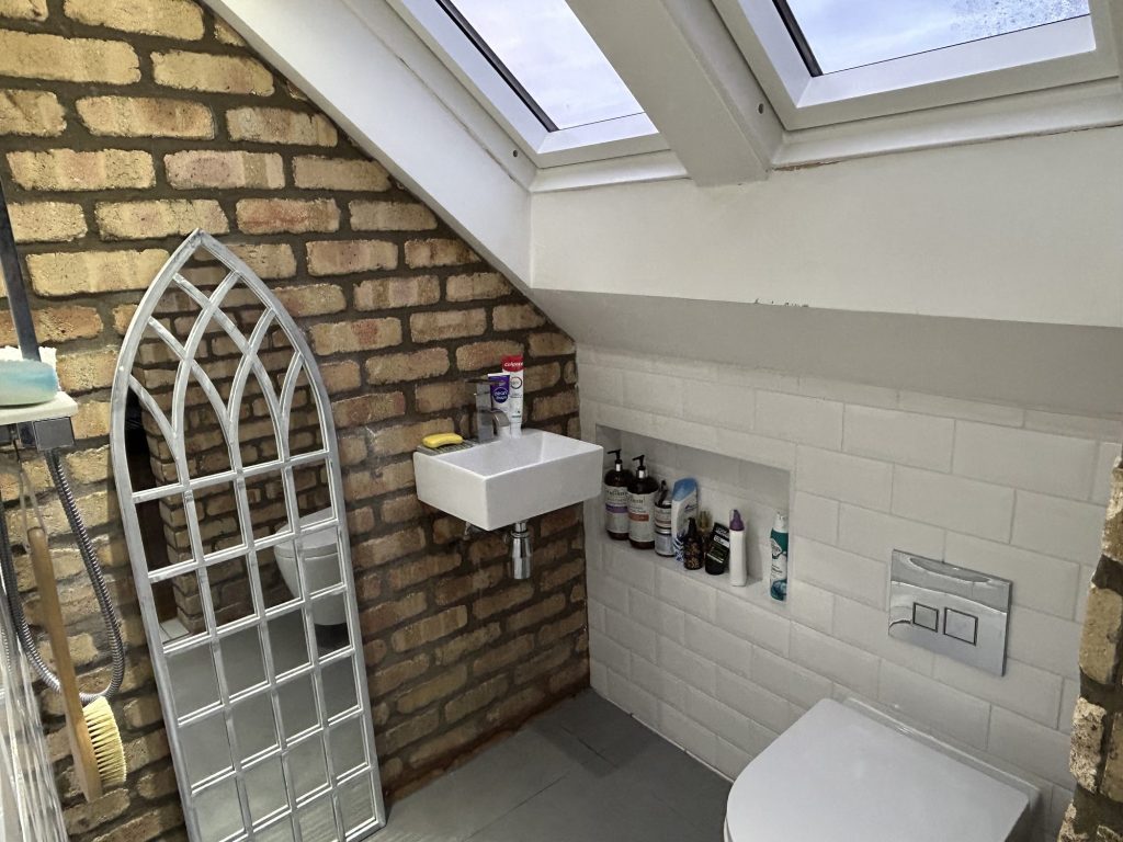Loft shower room with an exposed brick wall, white metro tiling, a wall-hung basin and WC, two Velux roof windows and a tall arched mirror.